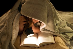 A young girl reading a book under the covers with a flashlight